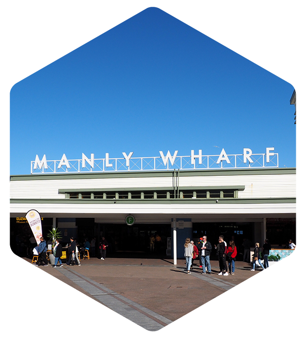 Building entrance with large white letters spelling 'MANLY WHARF' against a clear blue sky and people gathered in front.