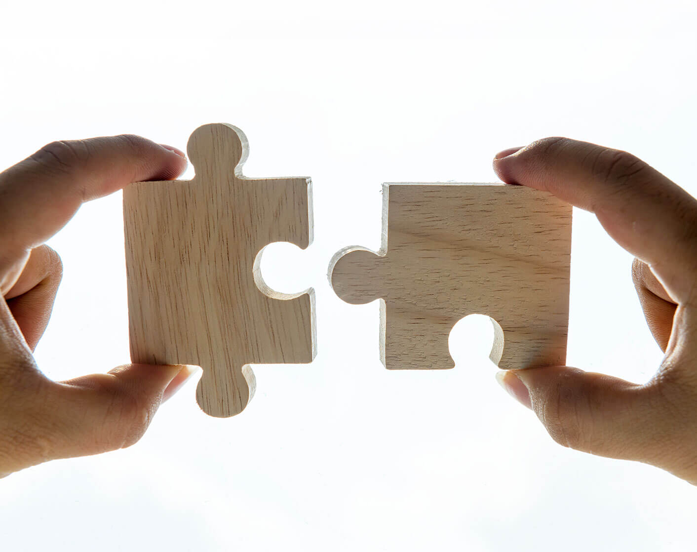 Two hands bringing two wooden puzzle pieces together against a bright white background.