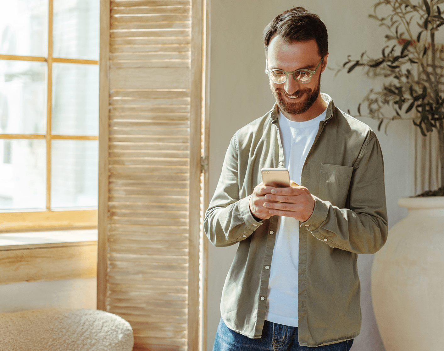 Smiling man with glasses holding a smartphone in a brightly lit indoor space with natural wood tones.