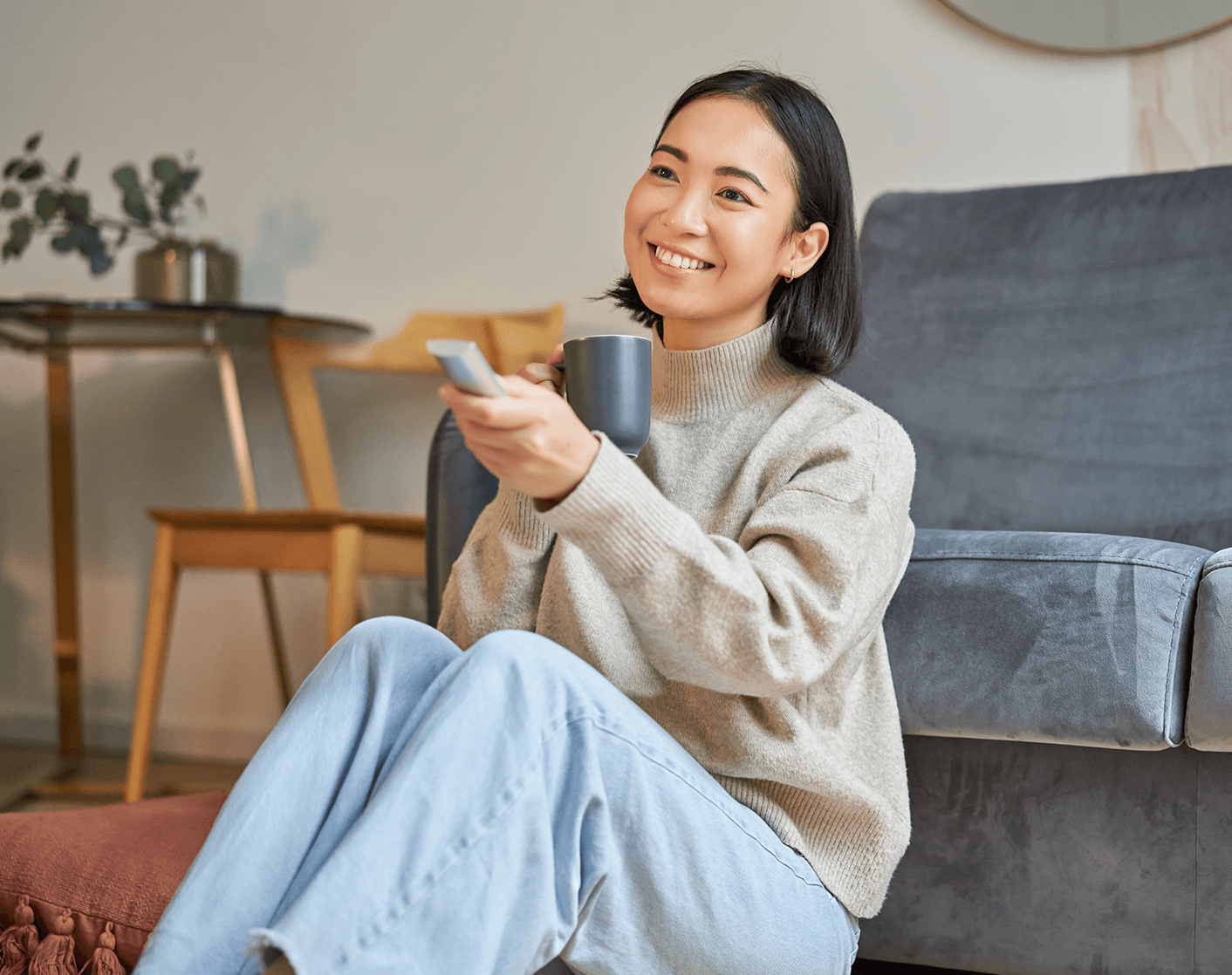 Woman relaxing on the floor next to a couch, holding a mug and pointing a remote control.