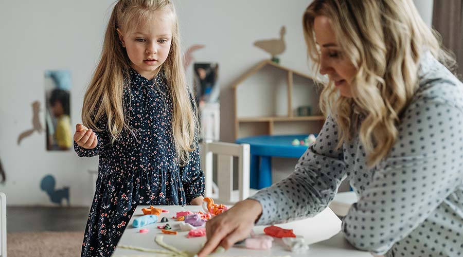 A parent plays with playdough in a preschool classroom. A young girl looks on.