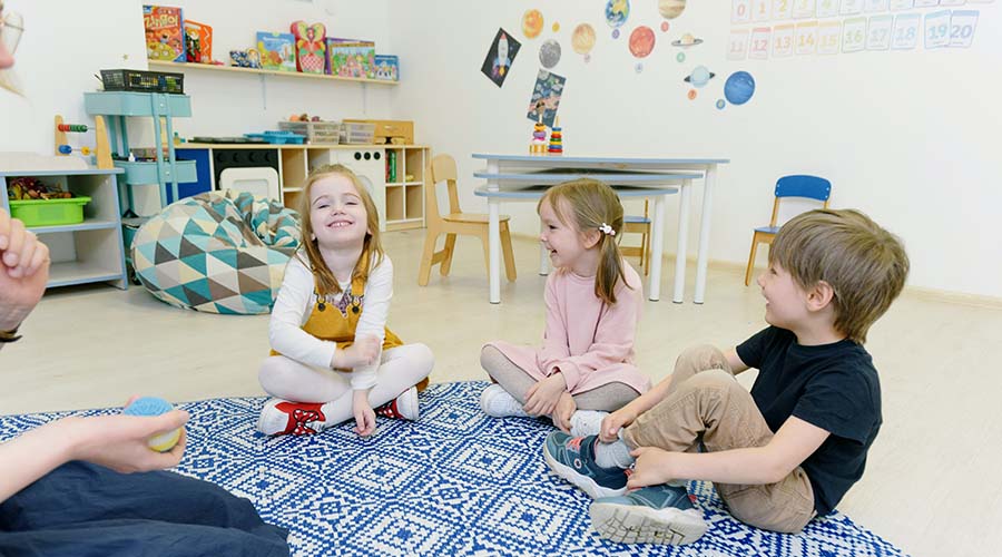 Three children are sitting cross-legged on a carpet in a child care classroom. They are smiling.