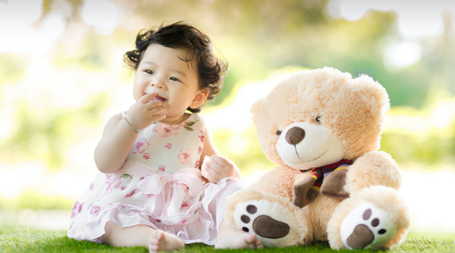 A young toddler sits with a teddy in a nursery garden