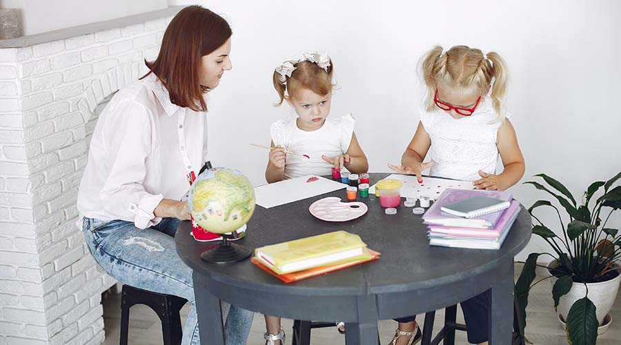 An early years teacher sitting with 2 girls, painting, in a nursery setting.