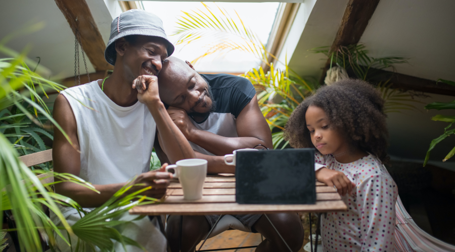 A photograph of two fathers drinking from cups and watching an iPad with their daughter.