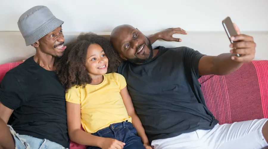 A photograph of a young child, posing for a selfie with her two fathers