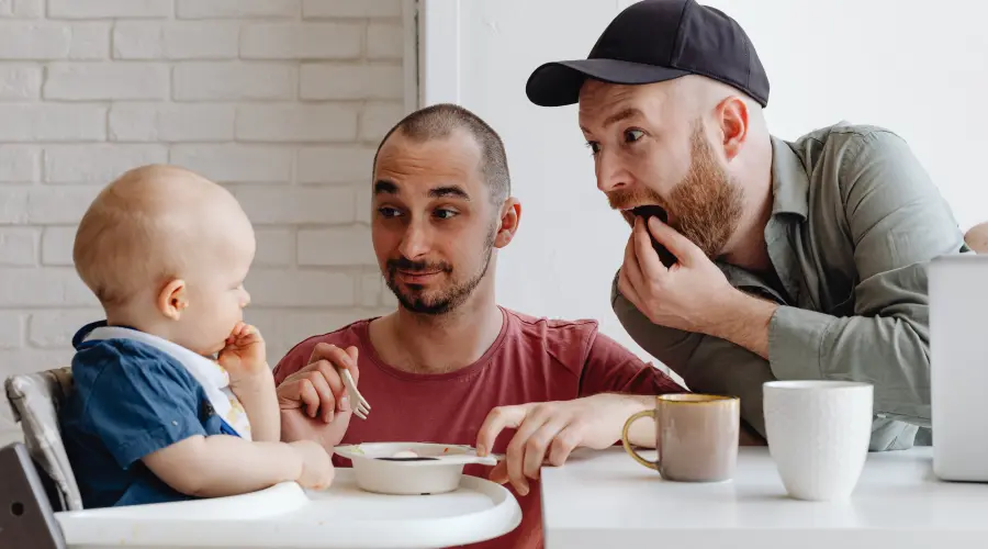 A photoraph of a young toddler, eating dinner with his two fathers