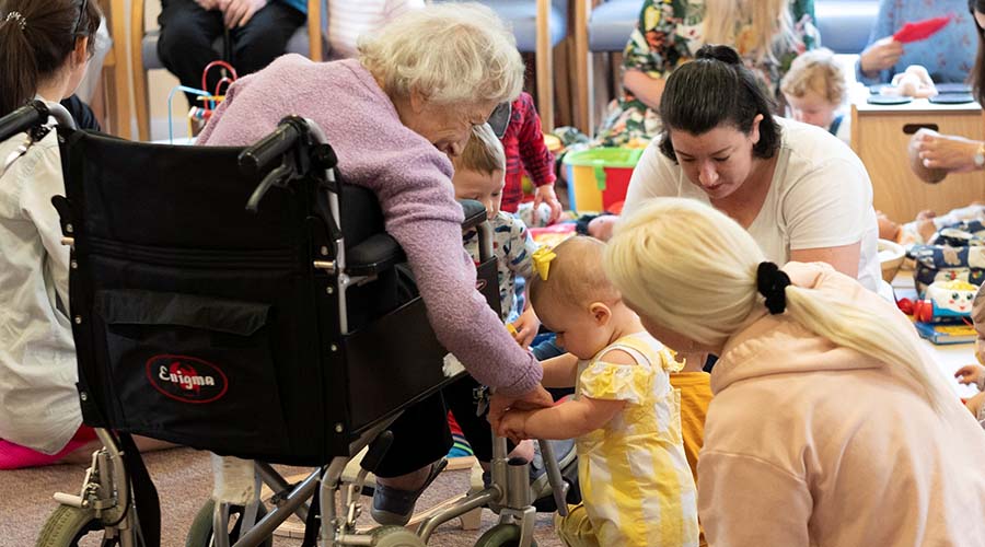 An elderly resident of Nightingale reaches down from her wheelchair to hold the hand of a baby, who is standing next to her.
