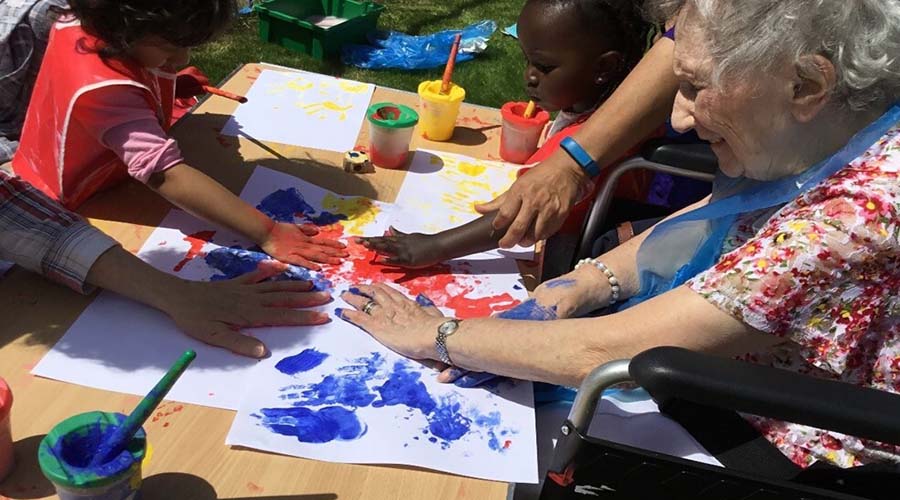 An older lady in a wheelchair is finger painting with two young children at a table, in a garden