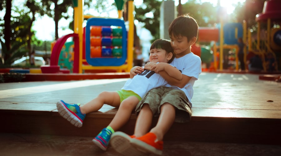 Two early years children, one younger, one older, hug together in a brightly-coloured nursery playground
