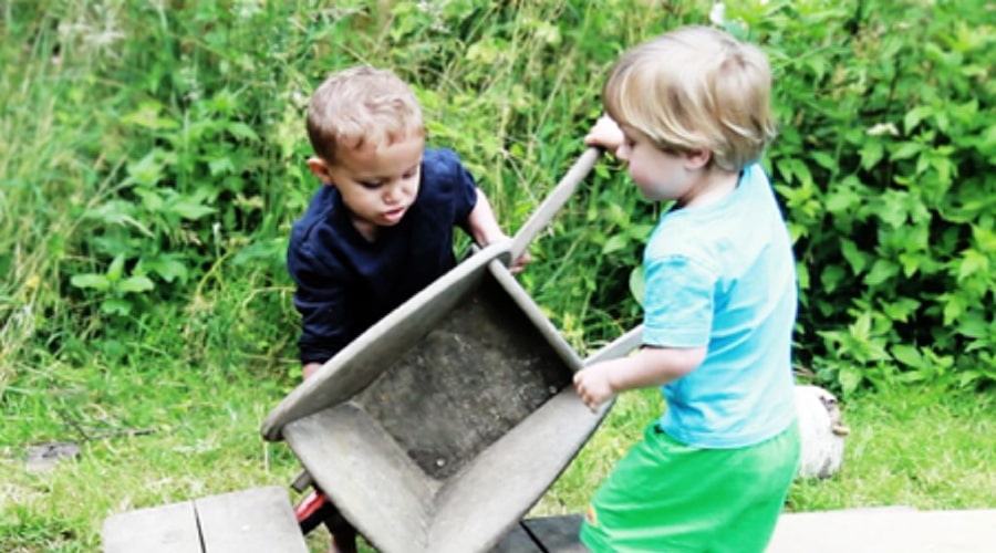 Two early years children work together to manoeuvre a wheelbarrow in a nursery garden.