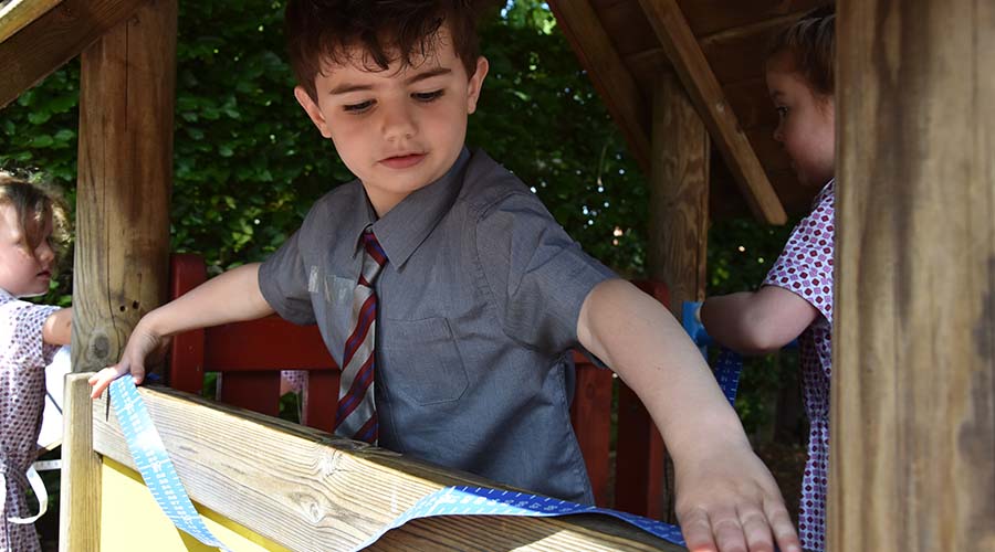 A little boy in a shirt and tie uses a tape measure inside a wooden playhouse.