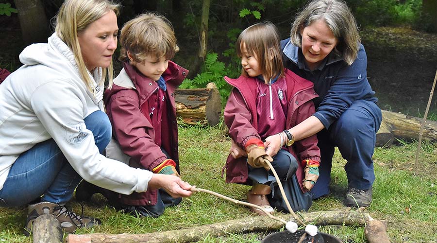 Two early years teachers support two children to roast marshmallows over a fire