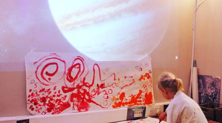 A photograph of a pre-schooler at Little Barn Owls nursery painting in red and orange with an image of Jupiter projected above them