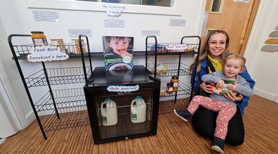 A photo of katie, and Early Years educator, with a young toddler, in front of the community pantry. There is a fridge in the centre and baskets either side of it. One is labelled "Food bank donations"