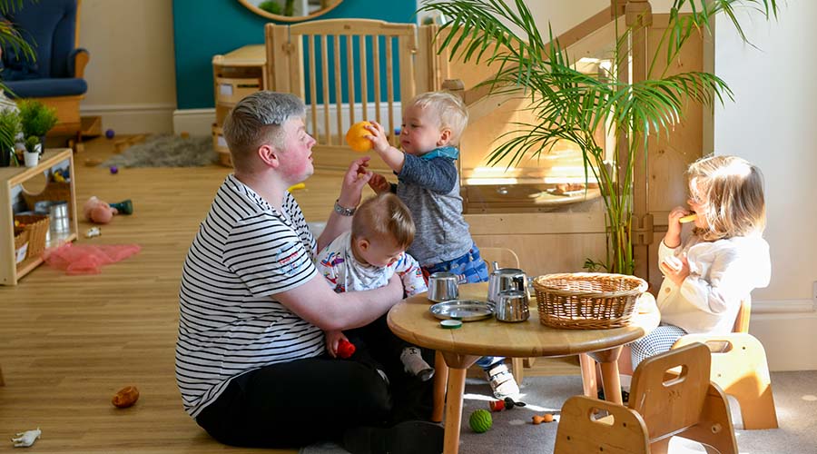 An Early Years educator sits at a small, wooden table with three toddlers. On of the little boys is sitting on the educator's lap, another is handing him a toy orange. A little girl pretends to eat a toy bun.