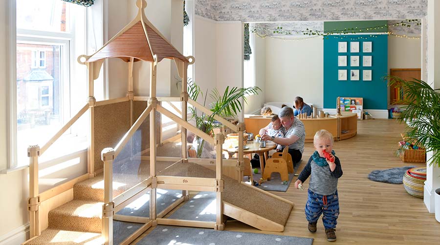 The toddler room at Kinder Park Nursery. A large wooden slide can with a toddler walking by it. There are two educators in the background playing with some other children.
