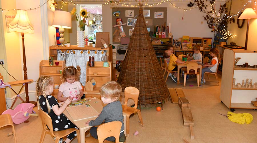 The preschool room at Kinder Park. 7 children sit at two wooden tables, with a wicker tent between them. the room is softly lit with lamps and fairy lights. There are wooden toys on the shelves and a tree in the back corner.