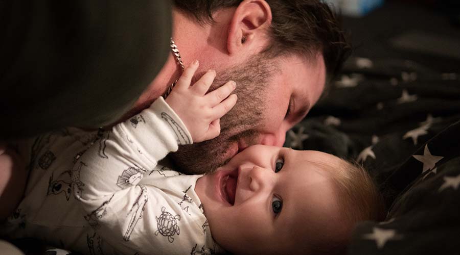 A father kisses his young baby on the cheek. The baby is smiling and holding the father's face.