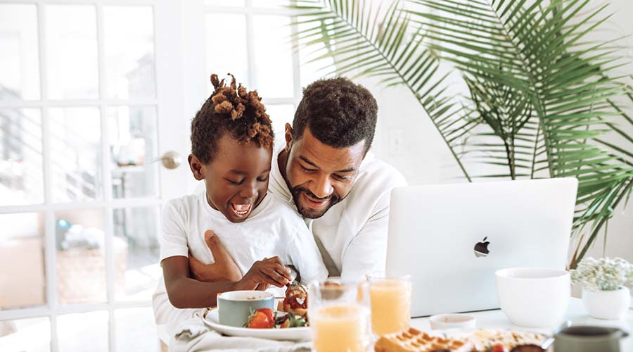 A father enters information for his child's nursery on a laptop. The son is sitting on the father's lap at a table, which also has breakfast foods on it.