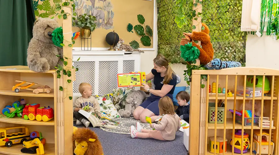 An early years educator reads to three toddlers in a cosy, jungle-themed book corner.
