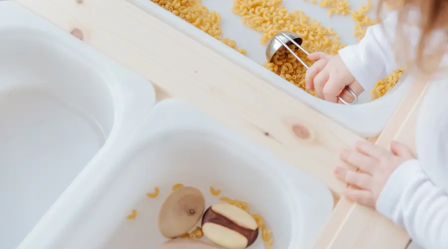 A toddler plays in a sensory tray filled with macaroni