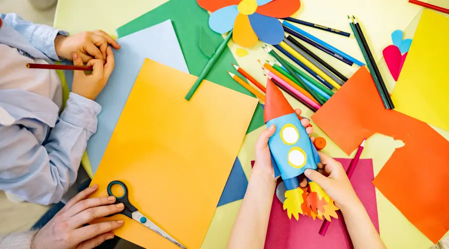 An overhead view of a craft table at a preschool or nursery. You can see two young children's hands and an adults' in between them. The table is covered in paper, pencils, and some finished crafts.
