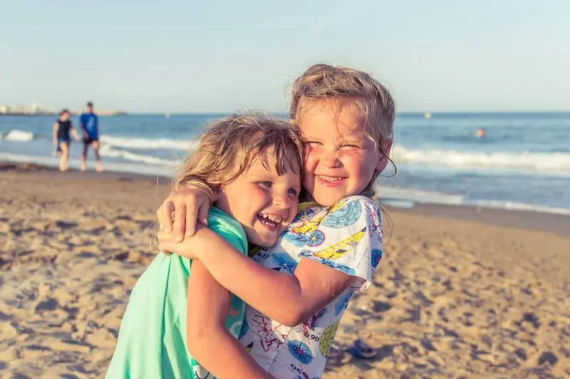 Two little girls hug. They are smiling on a beach.