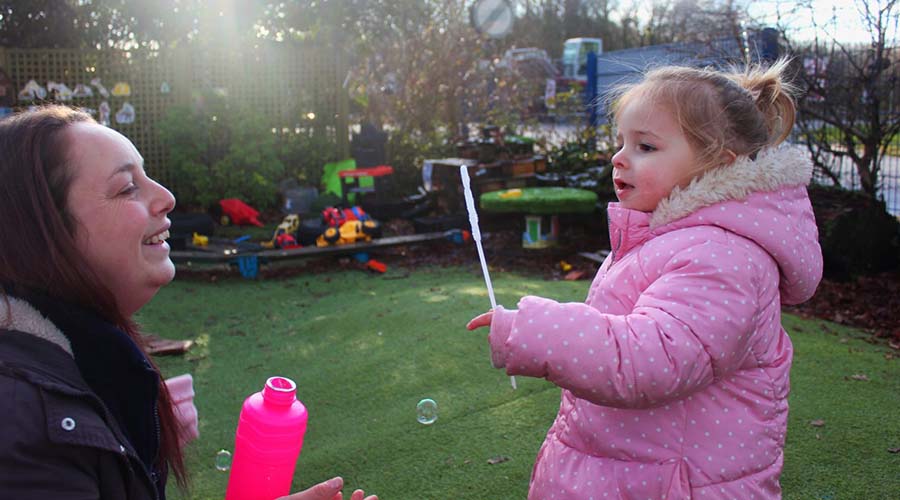 An Early Years teacher and a child in a pink coat blow bubbles together in a garden.