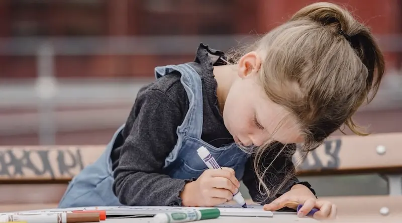 A photo of a preschool child holding a felt-tip pen, drawing in a book. 