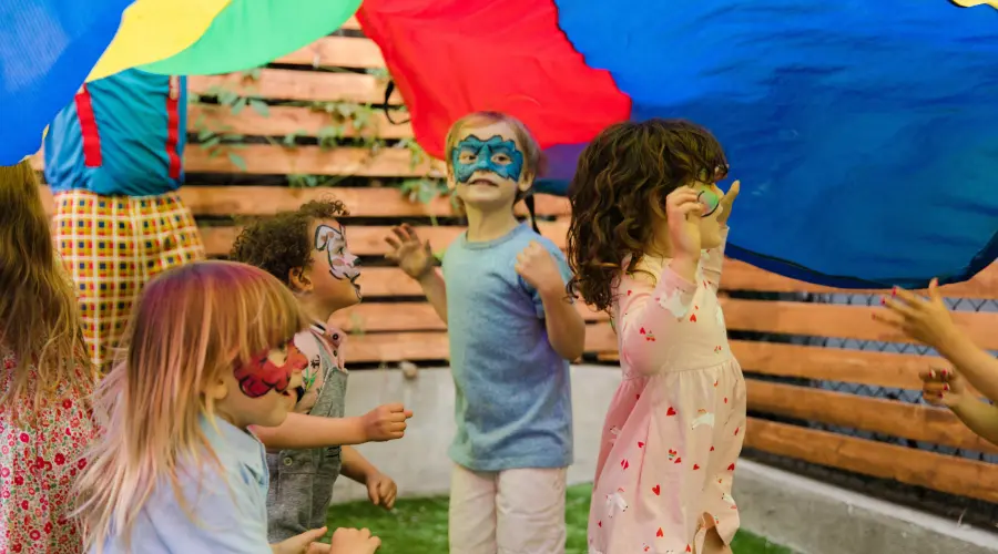 Several preschool children with face paint on play under a large colourful parachute