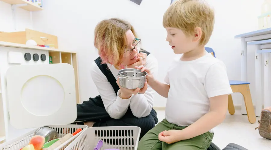 A parent plays in the home corner with a preschool-aged child. She is handing him a colander from a toy kitchen cupboard.