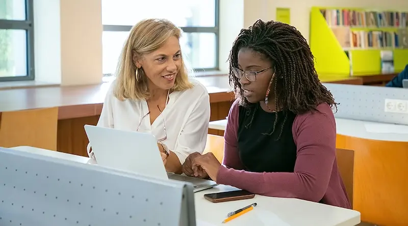 An Early Years apprentice and her college tutor sit together at a desk in the library. They are looking at an open laptop together.