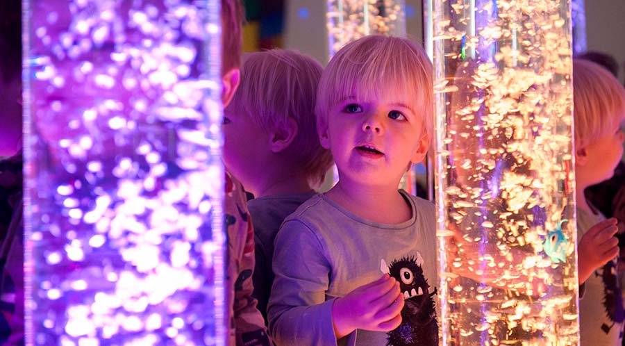 A little blonde boy sits among lit up bubble tubes in a sensory room.
