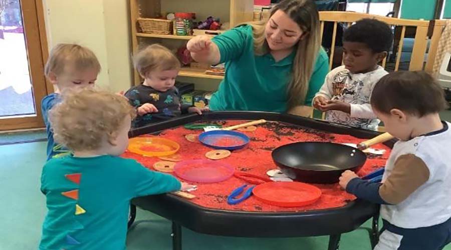 An early years educator kneels next to a Tuff Tray full of red sand. There are 5 toddlers around the tray, using the plates and tools to play with the sand.