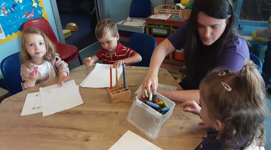 An early years educator sits at a table with three children. They are using pencils and wax crayons to make marks on paper.