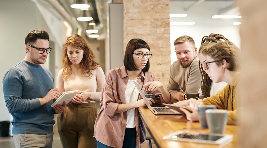 A group of adults in a meeting. A man and a woman looks at a notebook together and 4 adults stand around a laptop, talking together.