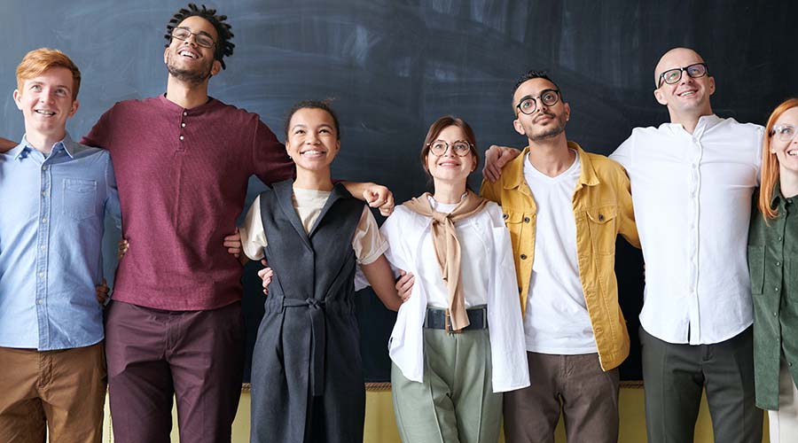 A group of teachers stand in front of a chalk board. They are arm-in-arm and smiling.