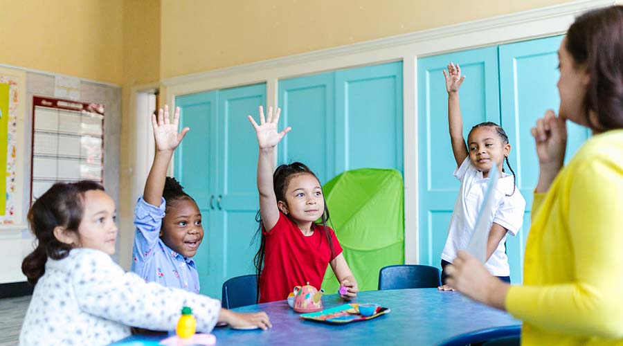 4 Early Years children are at a table, facing an educator. Three of them have one hand in the air.