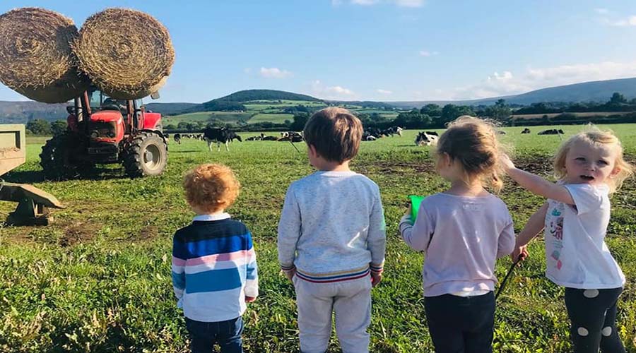 4 children are in a field, looking towards some cows and a tractor holding up two huge hay bales.