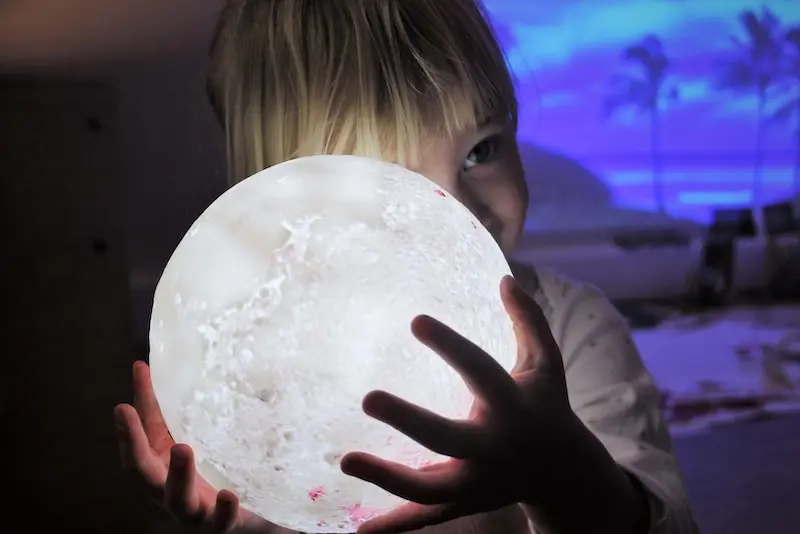 A child holds a glowing moon lamp, casting soft light on their face. The background shows a serene seascape with palm trees, evoking wonder and curiosity.