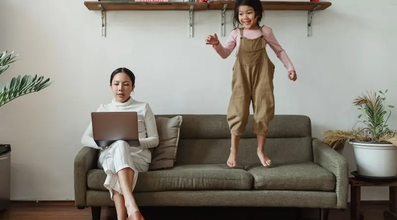 Mother working on a lap-top on a couch, while her daughter is jumping next to her