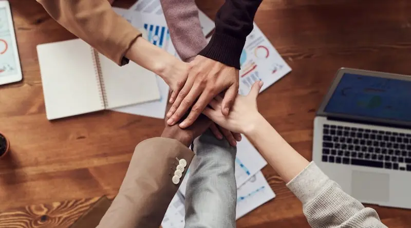 Hands crossing together in a team event over a busy board with papers and laptop