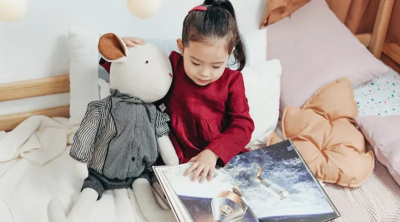 Little girl sitting with a plush toy reads a book