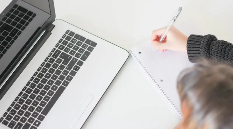 Person writing on paper next to a MacBook