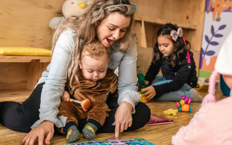 an teacher with an infant and a toddler behind her in a classroom