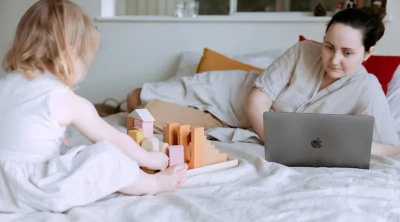 a girl playing with blocks while her mom checks her laptop on her bed