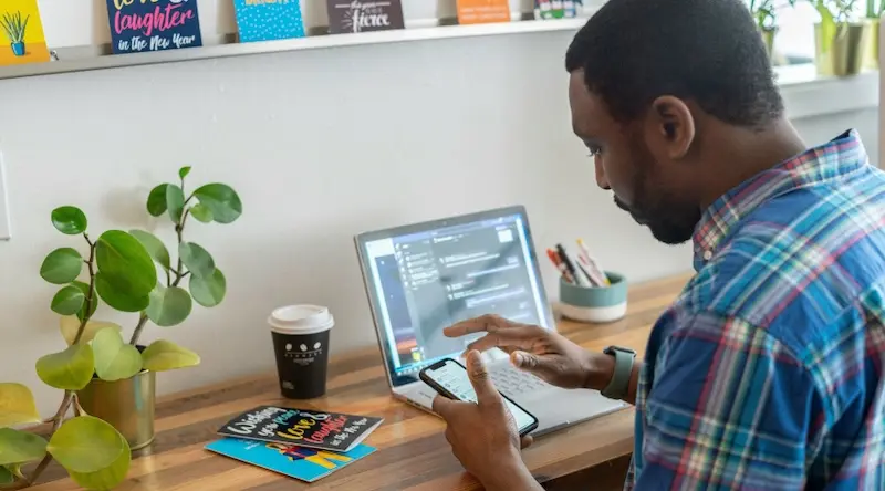 man at his desk on his laptop and iphone, books on wall