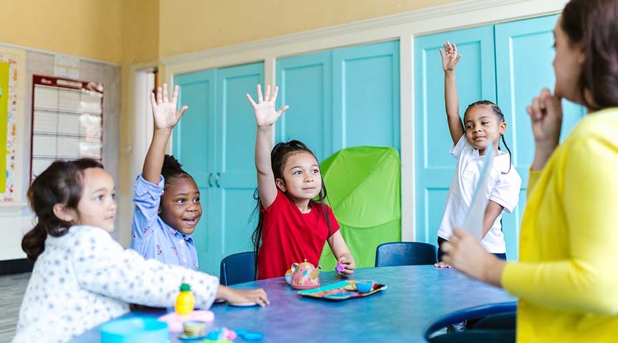 Several young children sit at a table, raising their hands to answer a question