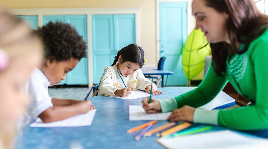 Children sit at a table with their teacher, drawing with colored pencils